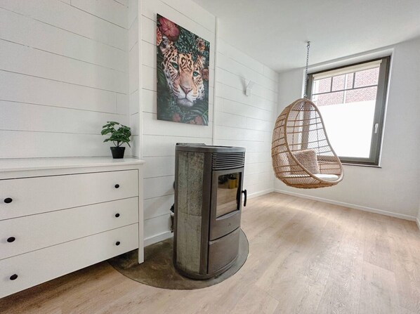 Een kamer met een witte muur, een luipaard schilderij, een hangstoel, een houtkachel en een raam.