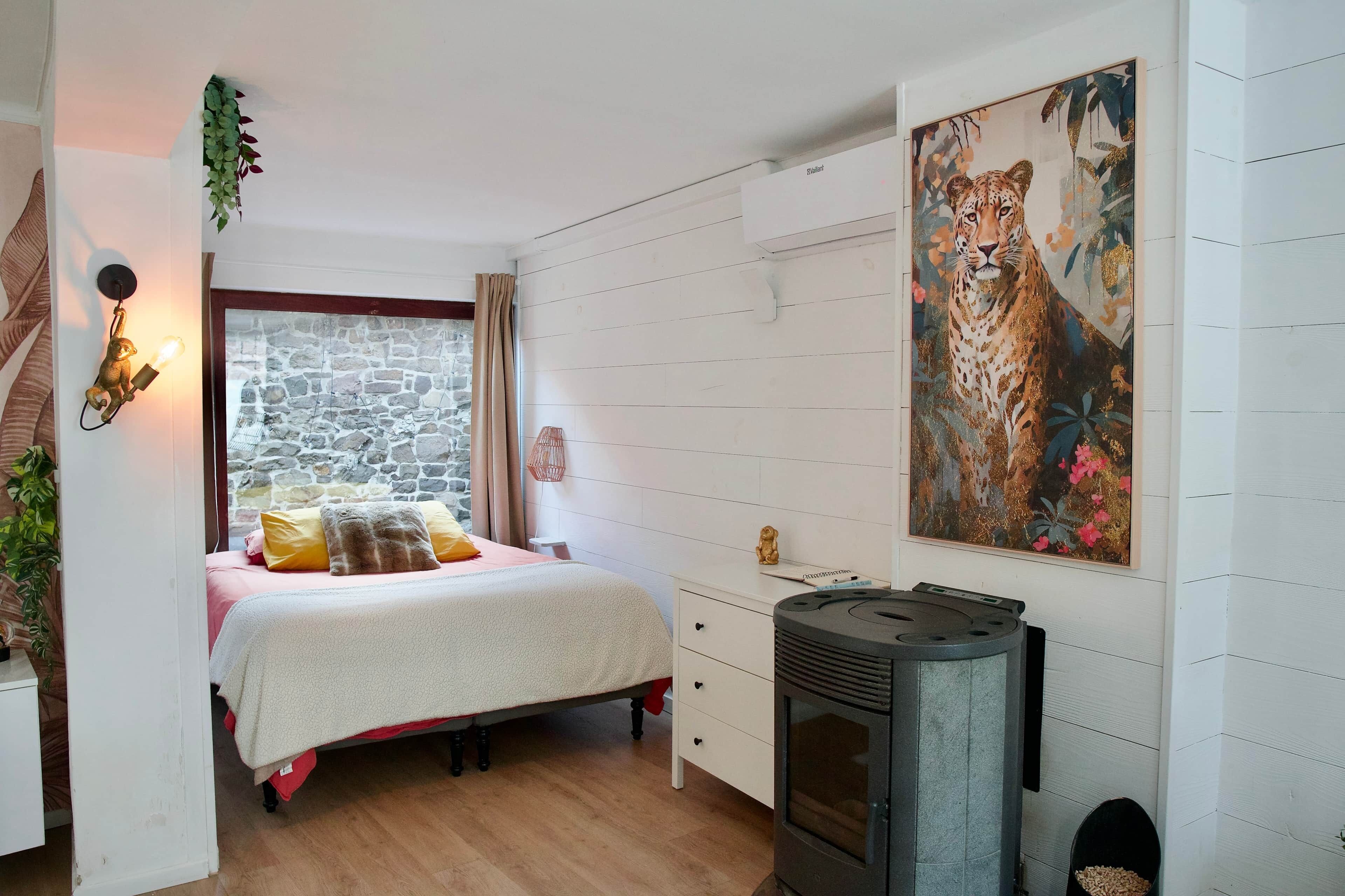 Une chambre avec un mur en pierre, un lit doté d’une tête de lit rouge, un grand tableau représentant un léopard et un poêle à bois.