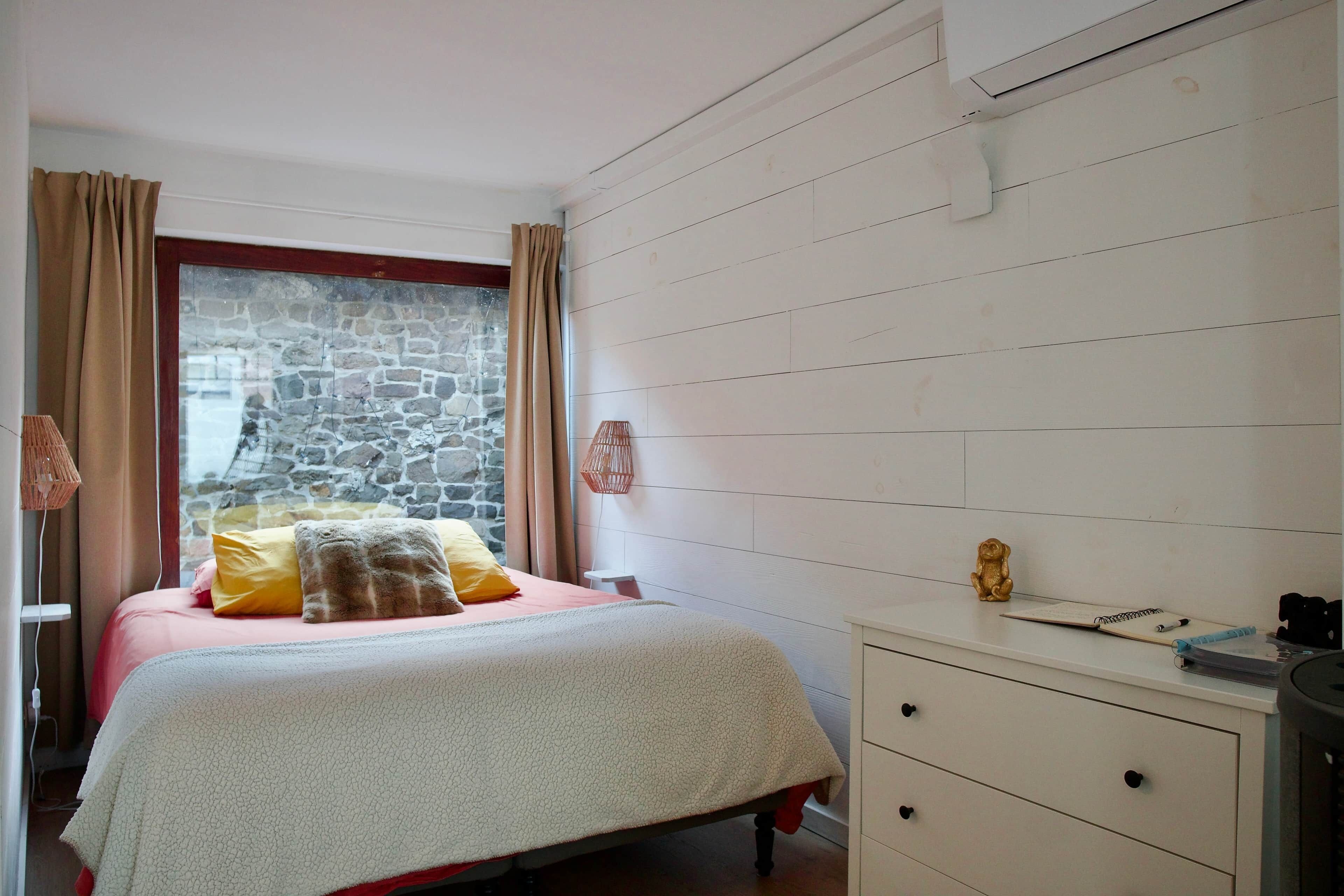 A bedroom with a stone wall, a bed with a white cover, a wooden dresser, and a small statue on the dresser.