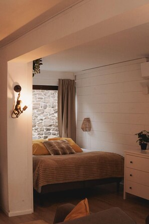 Een slaapkamer met een stenen muur, een bed met een gestreept deken, een bedlampje en een tafeltje met een plant.