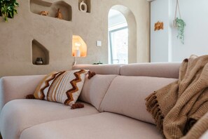 Een beige bank met een gekleurd kussen en een plaid, een wandplank met een beeldje, en een hangende plant.