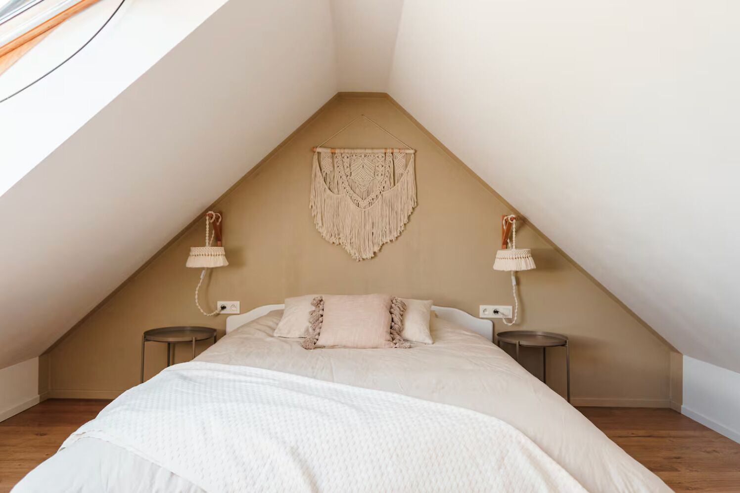 Ein Schlafzimmer mit schrägem Dach, einem Bett mit weißer Bettdecke, zwei Nachttischlampen und einem Wandbehang aus Makramee.