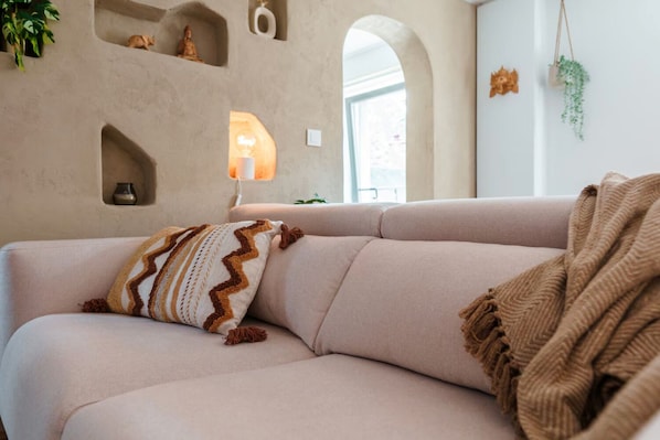 A beige sofa with a patterned pillow and a throw blanket, a wall-mounted shelf with a statue, and a hanging plant. - Le Bali - Zen & serenity (Profondeville)