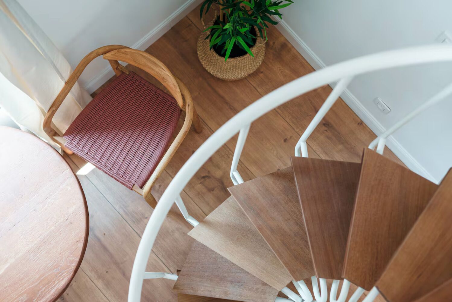A spiral staircase with wooden steps and a white railing, a wicker chair with a red cushion, and a potted plant on the floor.