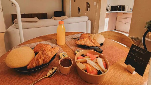Een ontbijttafel met croissants, een schaal fruit, een fles sap en een bordje met een welkomstbericht voor gasten.