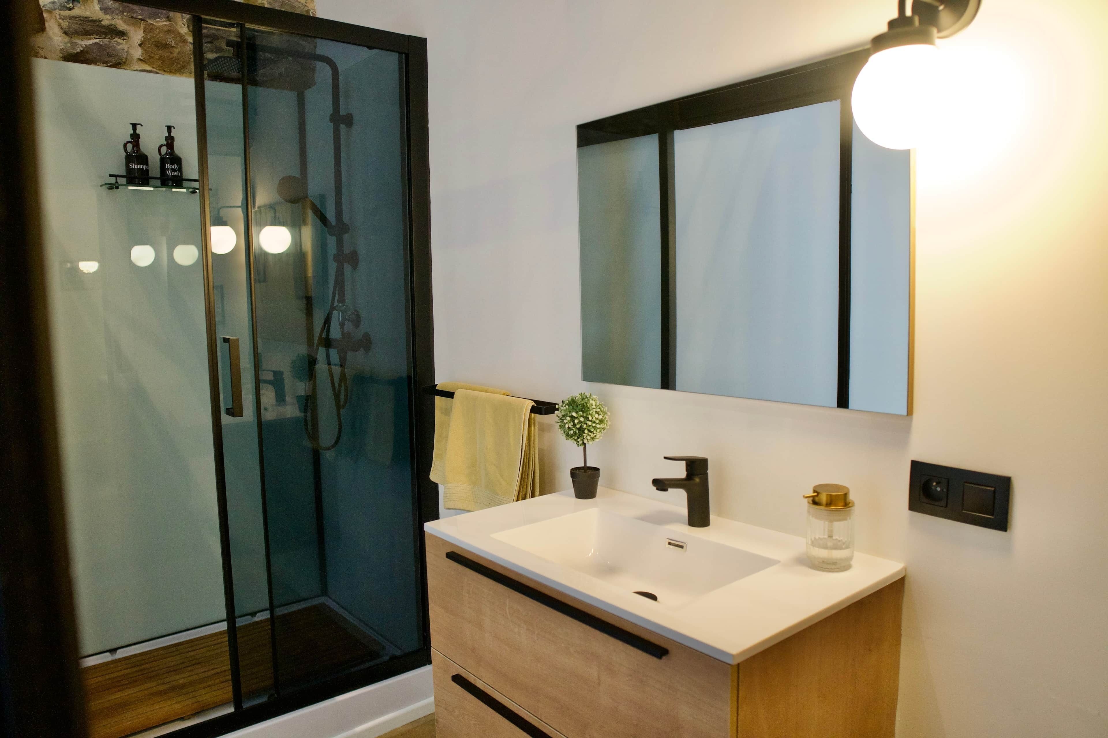 A modern bathroom with a glass shower enclosure, a white sink with a black faucet, and a mirror with a light above it.