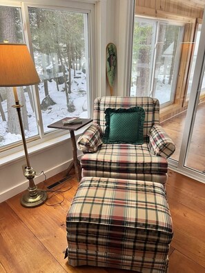 A cozy room with a plaid armchair and ottoman, a floor lamp, and a window with a view of a snowy landscape. - Dam Good Times - 90s Lake Getaway (Stoddard)