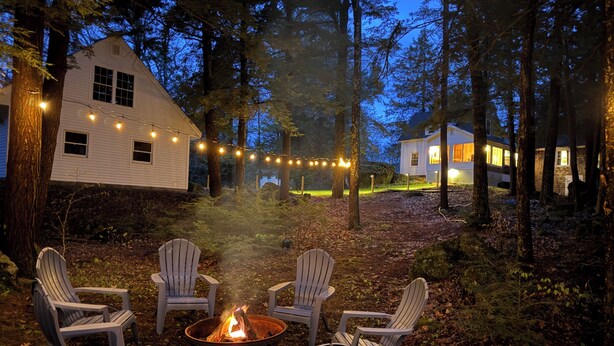 Un espacio al aire libre acogedor con sillas Adirondack alrededor de un brasero, rodeado de árboles y casas.