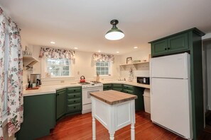 A kitchen with green cabinets, a white refrigerator, a wooden table, and a window with floral curtains. - Dam Good Times - 90s Lake Getaway (Stoddard)
