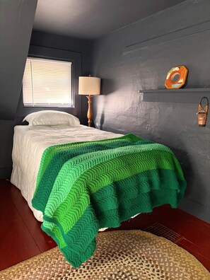A bedroom with a bed, a window with blinds, a lamp, and a shelf with decorative items. - Dam Good Times - 90s Lake Getaway (Stoddard)