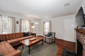 A living room with a brown leather sofa, a plaid armchair, a wooden coffee table, and a fireplace. - Dam Good Times - 90s Lake Getaway (Stoddard)