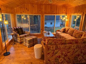 A cozy room with wooden walls and large windows, furnished with a sofa, a plaid ottoman, and a round table.