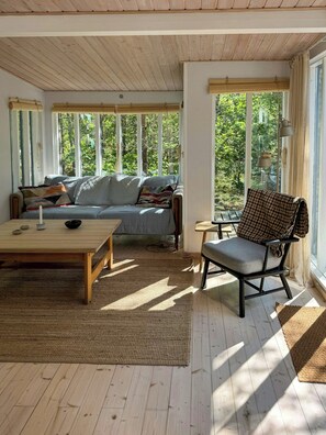A cozy room with a sofa, a chair, a table, and large windows offering a view of trees. - Forest house near Bakka Harbor and Kadeau (Aakirkeby)