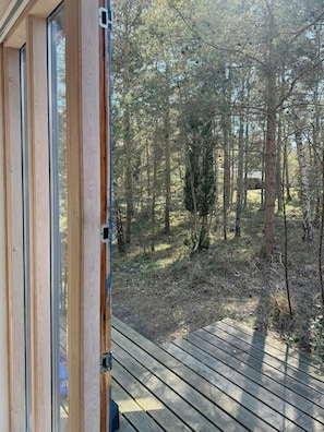 Eine Holzterrasse führt zu einem bewaldeten Gebiet, durch eine Glastür ist die Aussicht sichtbar.