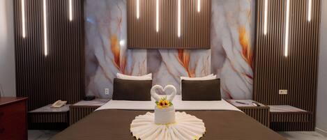 A hotel room with a large bed, a heart-shaped towel, a vase with a flower, and a marble-patterned wall.