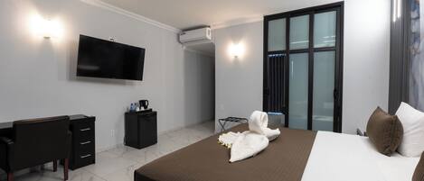 Una habitación de hotel moderna con una cama grande, un televisor de pantalla plana en la pared, un escritorio con cajones, un mueble pequeño y un cerramiento de vidrio corredera que da a un balcón.