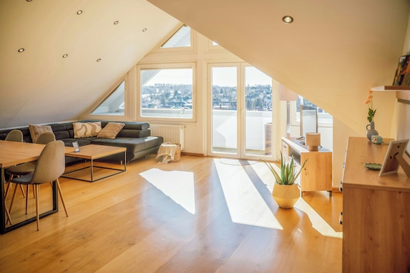 Ein modernes Loft mit schrägem Dach, einem Essbereich, einer Couch, einem Couchtisch, einer Topfpflanze und Blick auf die Stadt.