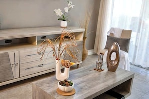 Ein modernes Wohnzimmer mit Couchtisch aus Holz, einer weißen Vase mit Trockenpflanzen, einer kleinen weißen Schale, einer dekorativen Metallskulptur und einem gepolsterten Sessel.