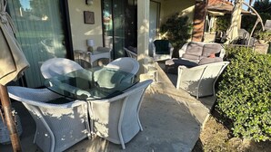 Eine Terrasse mit Korbmöbeln, einem Glastisch und Blick auf ein Haus mit Vordach und Palmen.