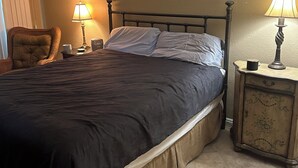 Ein Schlafzimmer mit einem Bett, zwei Nachttischen mit Lampen, einem Sessel und einem Fenster mit Jalousien.