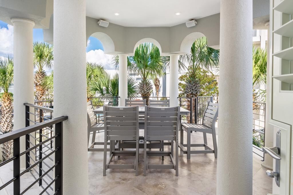 An outdoor patio with a table and chairs, surrounded by white columns and palm trees.