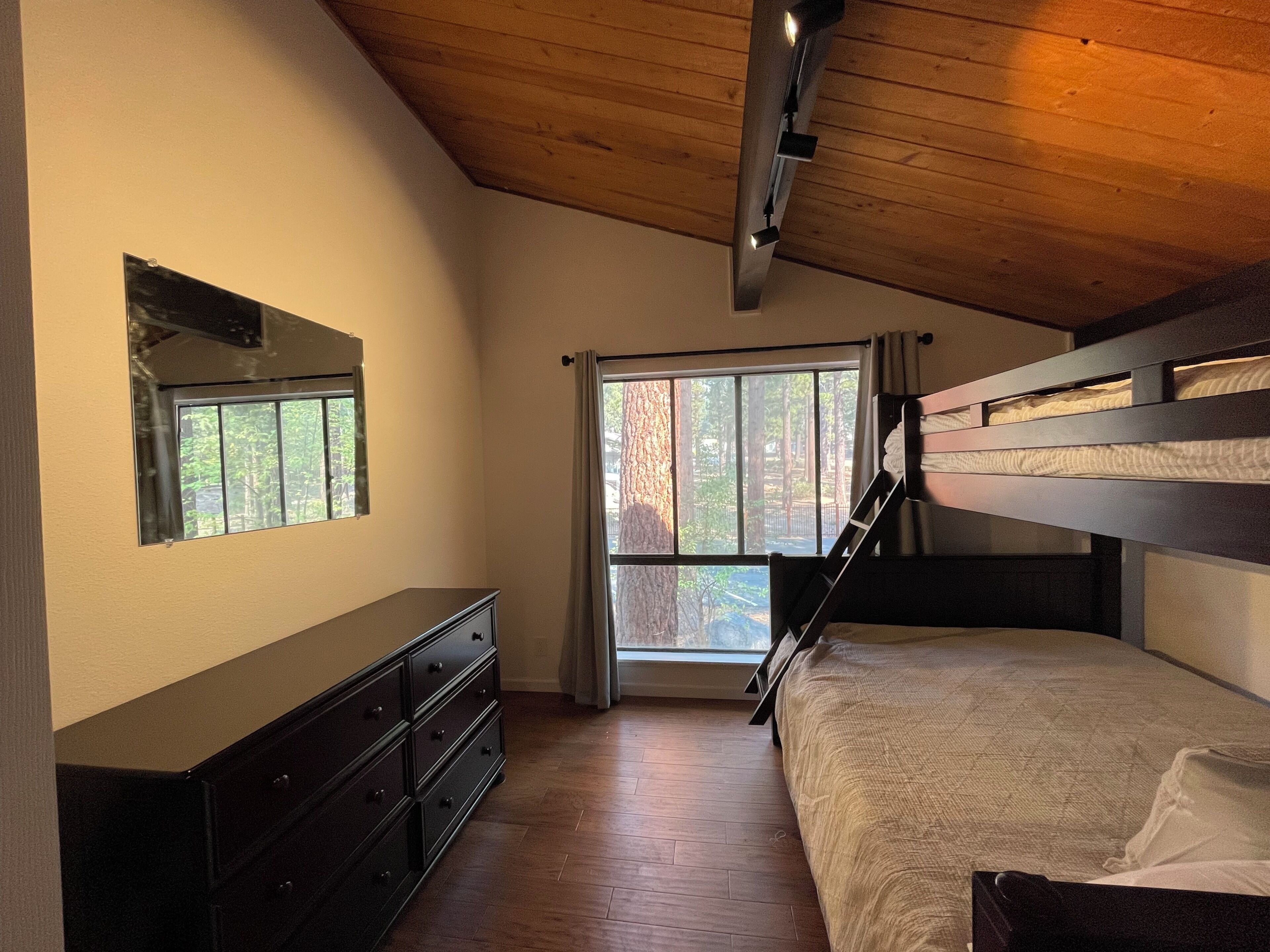 A bedroom with bunk beds, a dresser, a window with curtains, and a wooden ceiling.