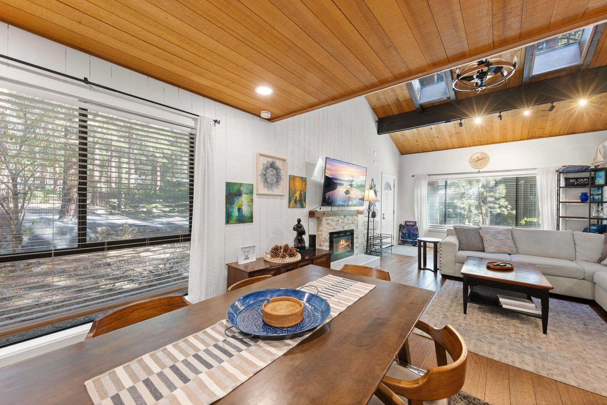 A spacious living area with a wooden ceiling, a fireplace, and a large window overlooking a forest.