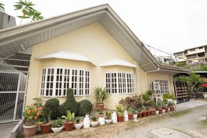 A residential building with a sloped roof, multiple windows with white frames, and a variety of potted plants in the front yard.