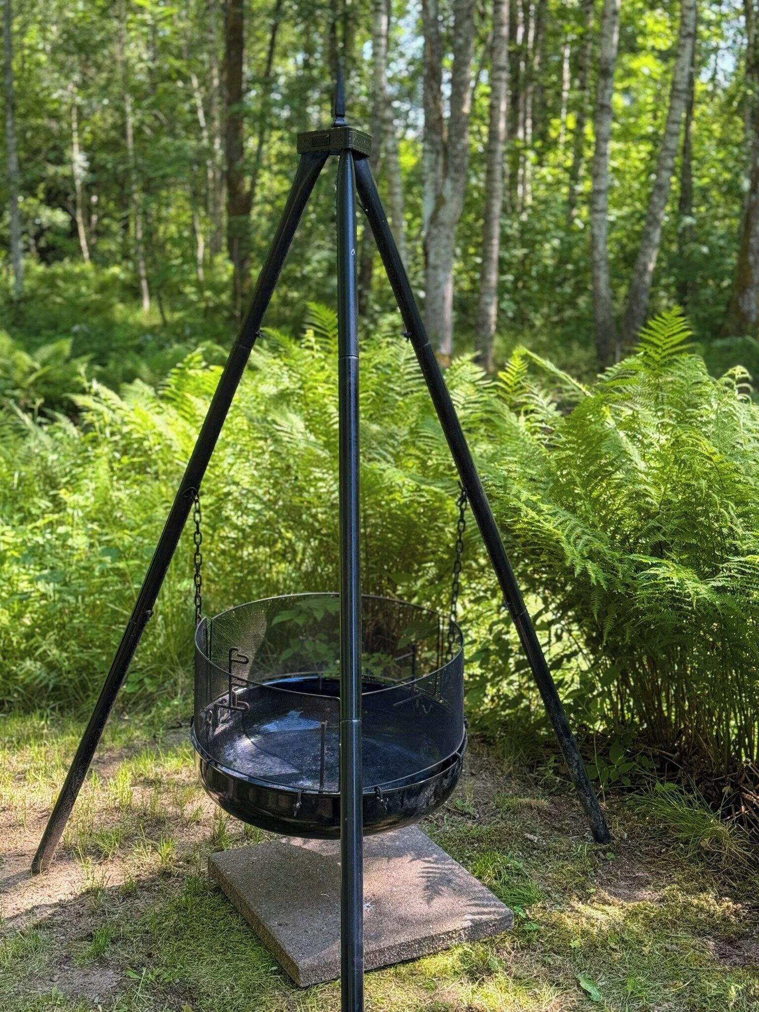 A metal hanging chair suspended by a tripod stand in a forested area.