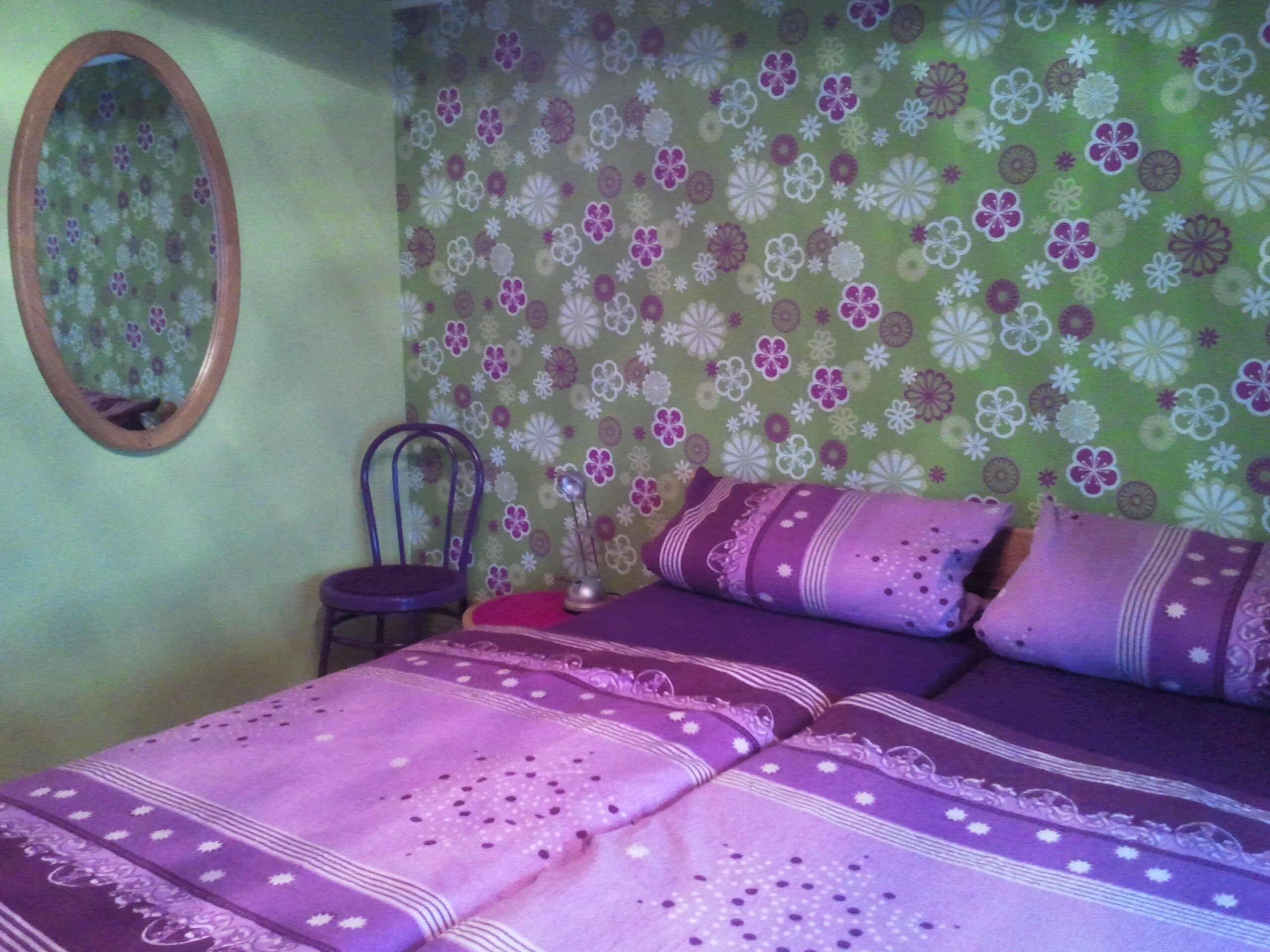 A bedroom with a floral wallpaper, a mirror, a chair, and a bed with purple bedding.