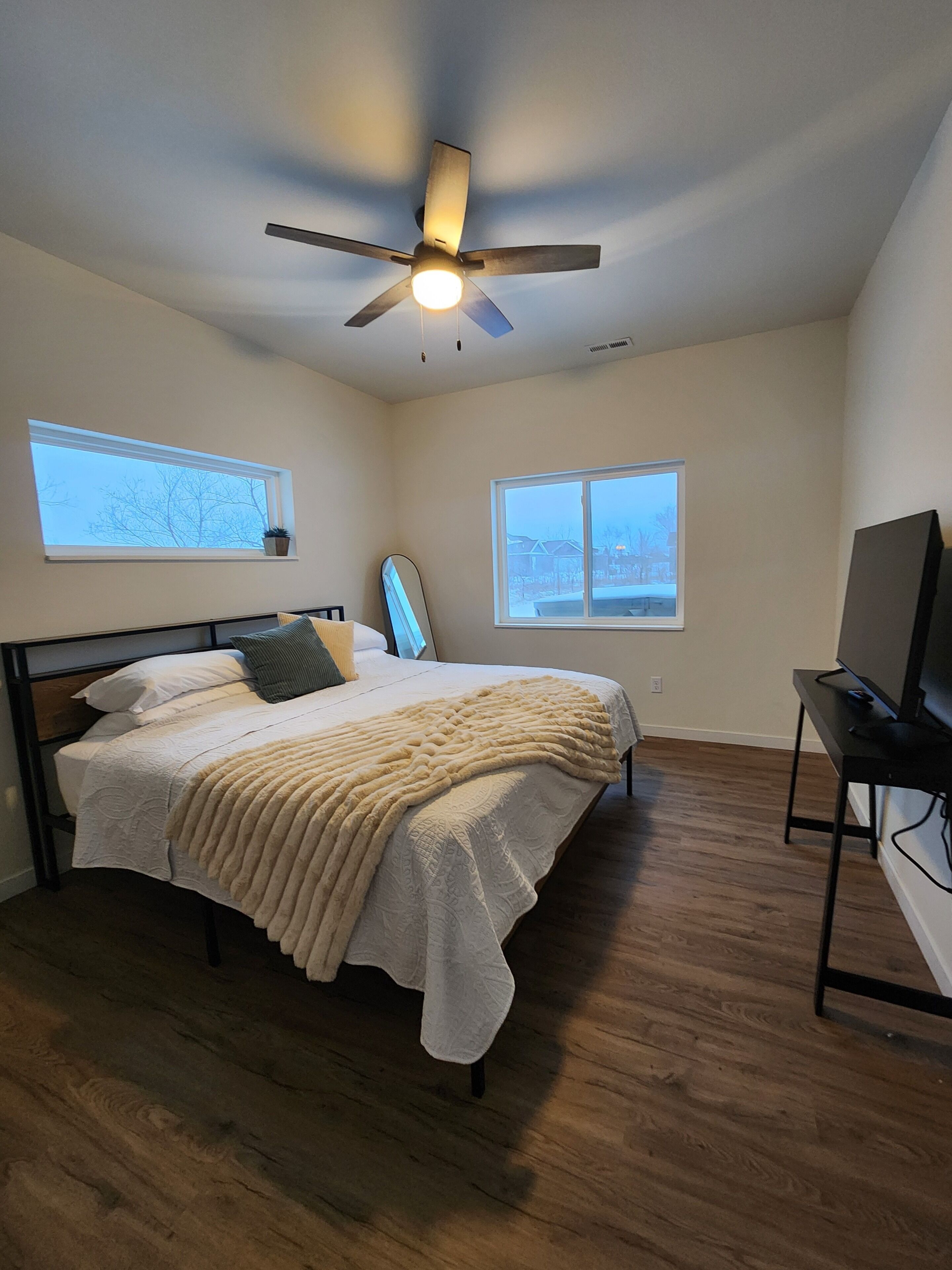 A bedroom with a bed, a ceiling fan, a TV stand, and two windows.