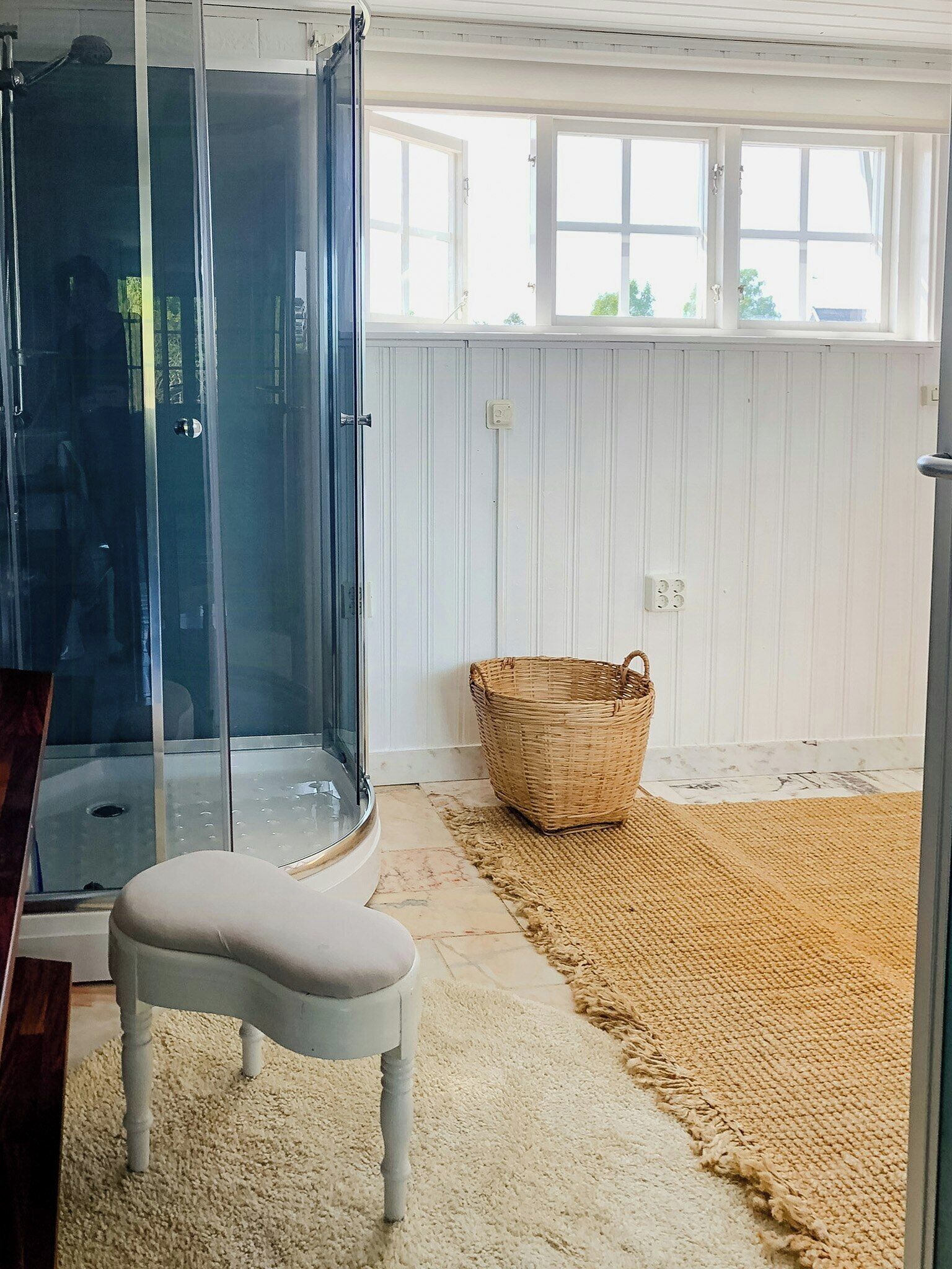 A room with a glass shower enclosure, a white cushioned bench, a woven basket, and a large window with white shutters.