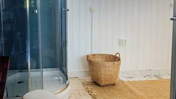 A room with a glass shower enclosure, a white cushioned bench, a woven basket, and a large window with white shutters.