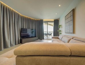 A modern living room with a large sofa, a flat-screen TV, and a balcony with a table and chairs. - Elegant Sea View 3BR Apartment in Qawra | Sleeps 6 (San Pawl il-Baħar)