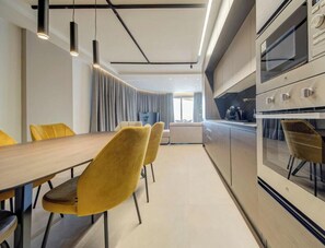A modern kitchen with a wooden dining table and yellow chairs, a built-in oven, and pendant lighting. - Elegant Sea View 3BR Apartment in Qawra | Sleeps 6 (San Pawl il-Baħar)
