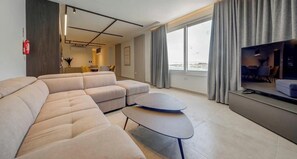 Ein modernes Wohnzimmer mit einer Eckcouch, einem kleinen runden Tisch, einem Flachbildfernseher und einem großen Fenster mit Vorhängen.