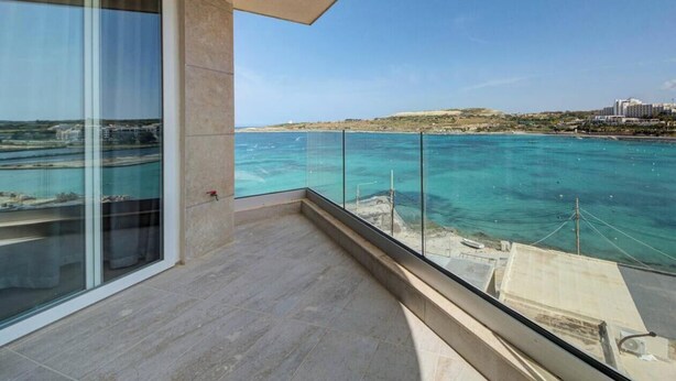 Ein Balkon mit Glasgeländer mit Blick auf eine Küstenlandschaft, blaues, klares Wasser und entfernte Gebäude.