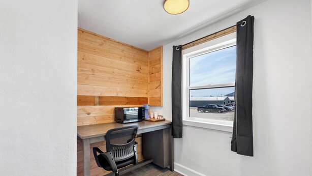 Un espacio de oficina pequeño con un escritorio de madera, una silla de oficina negra, un microondas y una ventana con cortinas negras.