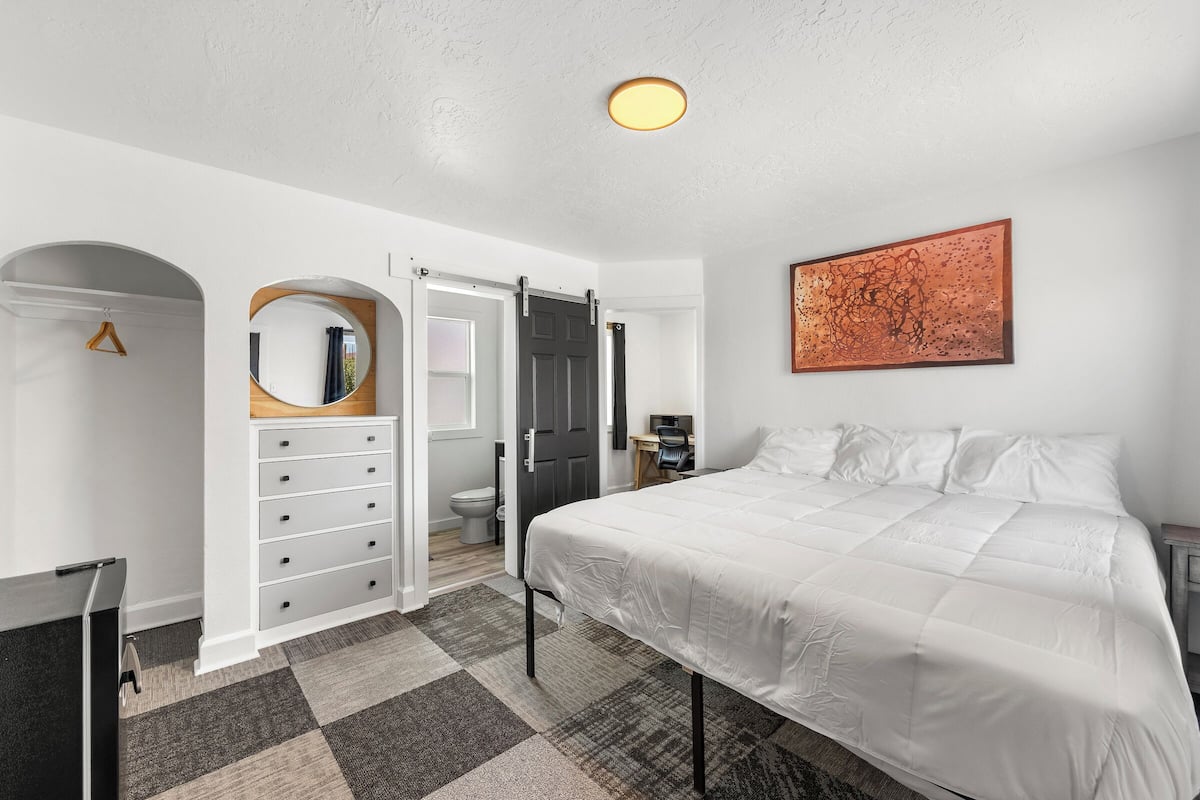 Een slaapkamer met een bed, een kapstok, een spiegel, een deur en een schilderij aan de muur.