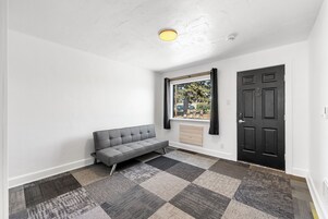 Una habitación con un sofá gris, un suelo de alfombra a cuadros, una puerta negra y una ventana con cortinas.