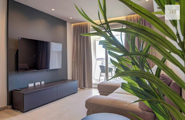 Ein modernes Wohnzimmer mit einem Flachbildfernseher an einer dunklen Wand, einer Couch, einem Couchtisch und einer großen Pflanze.
