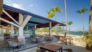 An outdoor seating area with tables and chairs, a covered section, and a view of the beach and ocean.