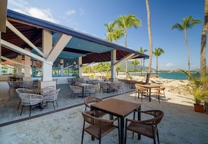 Un espace extérieur aménagé avec des tables et des chaises, une partie abritée et une vue sur la plage et l’océan.