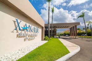 Una entrada moderna de hotel con un letrero que dice 'Xanadu Resort & Residences by Hodelpa', que cuenta con un césped cuidado y palmeras.