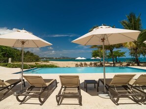 Zona de piscina con sillones y sombrillas, vista despejada a la playa y al océano, bajo un cielo azul tranquilo.