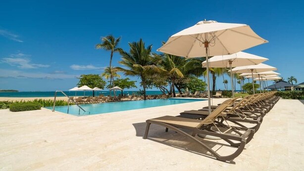 A poolside area with lounge chairs and umbrellas, palm trees, and a clear view of the ocean.