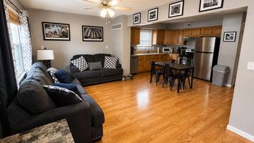 A living room with a sofa, a dining table, and chairs, a kitchen with wooden cabinets and appliances, and a ceiling fan.