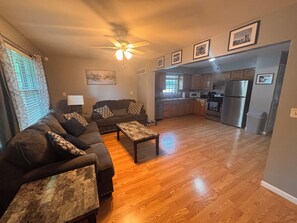 A living room with a sofa set, a coffee table, a ceiling fan, and a kitchen area with appliances. - KC Strong: Beautiful 2 BR Home, Close to stadiums and downtown. (Independence)