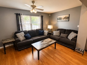 A living room with a sofa set, a coffee table, a floor lamp, a ceiling fan, and a window with curtains.