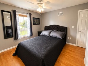 A bedroom with a bed, a ceiling fan, a window with curtains, a framed picture on the wall, and a door.